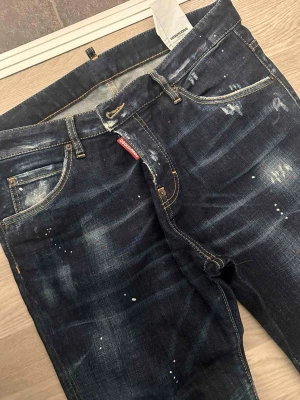 Mörkblå Dsquared2 jeans med slitningar original ÄKTA - Snygga mörkblå jeans från Dsquared2 med slitna detaljer, färgstänk och en röd logotagg vid fickan. Jeansen har klassisk femficksdesign, raka ben och kontrastsömmar. Perfekta för en trendig streetstil.