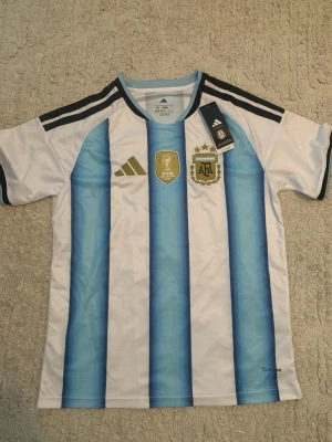 Argentina fotbollströja Adidas - Snygg Argentina fotbollströja från Adidas med klassiska blå och vita ränder, svarta detaljer och guldiga märken på bröstet. Tröjan har AFA-logga, FIFA-märke och tre stjärnor ovanför loggan. Tillverkad i lätt och ventilerande material.
