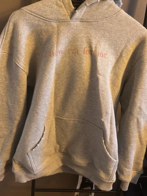 Grå hoodie med tryck framtill - En klassisk grå hoodie med huva och känguruficka framtill. Tryck i ljusrosa text på bröstet. Tillverkad i mjukt sweatshirtmaterial, perfekt för en avslappnad stil. Ribbstickade muddar vid ärmslut och nederkant.
