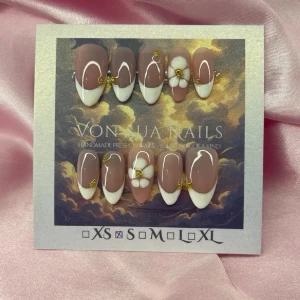 SoftBloom(S);Medium Almond - Snygga handgjorda press-on naglar i en medium mandelform. Designen har en nude bas med vit fransk tipp, vita swirl-detaljer och -blommor med gulddetaljer. Perfekt för dig som vill ha en trendig och elegant look. Kommer i S: 15 mm, 11,5 mm, 12 mm, 11 mm, 9 mm 