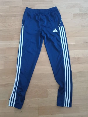 Blå Adidas träningsbyxor - Snygga blå träningsbyxor från Adidas med klassiska vita ränder längs benen och elastisk midja. Byxorna har smal passform och är perfekta för sport eller träning. Adidas-logga på ena benet och dragkedjor vid bensluten och fickor med dragkedjor.