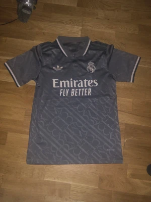 Real Madrid Adidas grå matchtröja - Ny Snygg grå Real Madrid matchtröja från Adidas med klubbmärke och Adidas-logga på bröstet. Tröjan har korta ärmar, krage med knapp och trycket 'Emirates Fly Better' på framsidan. Materialet är lätt och andas, perfekt för fotboll eller träning.