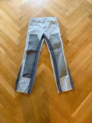 Patchwork jeans  - Unika patchwork jeans i ljusgrått och mörkgrått med raka ben och råa, fransiga benslut. Dekorerade med små nitar och håldetaljer på både fram- och baksida. Flera fickor och coola sömmar ger en trendig och avslappnad vibe. Perfekta för dig som gillar statement-plagg.
