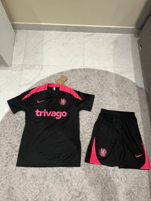 Chelsea svart och rosa fotbollskit Nike - Säljer ett svart Chelsea fotbollskit från Nike med rosa detaljer och tryck. Tröjan har rosa axelpartier, klubbmärke och sponsortryck, och shortsen har matchande rosa detaljer och klubbmärke. Perfekt för träning eller match!