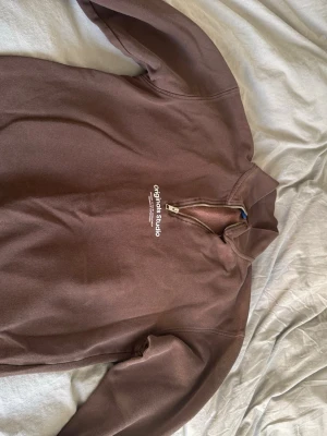 Brun quarterzip från Originals Studio - Säljer en brun quarterzip hoodie från Originals Studio med vit text på bröstet. Tröjan har hög krage, dragkedja och långa ärmar med muddar. Perfekt för en chill och stilren look. Materialet känns mjukt och skönt mot huden.