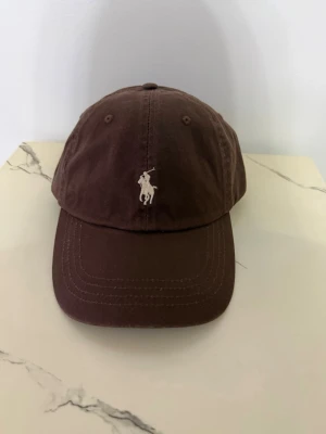 Brun keps från Polo Ralph Lauren - Snygg brun keps från Polo Ralph Lauren med klassisk broderad logga framtill och 'POLO' broderat baktill. Justerbar rem baktill för perfekt passform. Tillverkad i bomullstyg med böjd skärm, perfekt för en avslappnad och stilren look.