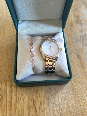 Roséguldklocka Ivy N Rox med armband - Snygg roséguldklocka från Ivy N Rox med länkarmband och gnistrande stenar runt urtavlan. Urtavlan är vit med eleganta detaljer och medföljande armband har hjärtformade länkar. Perfekt accessoar för att lyfta din stil. Kommer i orginalasken. Inga defekter, oanvänd