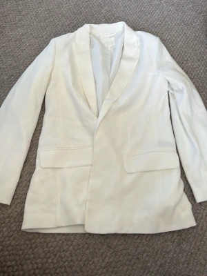Vit kavaj med klassisk passform - Stilren vit kavaj med lång ärm och klassisk krage. Kavajen har två framfickor med lock och är tillverkad i ett slätt material som ger en elegant look. Perfekt för dig som vill ha en enkel och snygg kavaj till garderoben.
