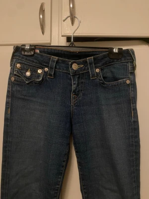 Mörkblå jeans från True Religion - Säljer mina fina true religion jeans, därav att jag använder dem för få gånger. De är i jättebra skick, förutom de små slitningarna längst ner på kanten av benen (bild 6 &7). Midjemåttet är ca 38 cm tvärs över och innebenslängden är 80 cm. Jag skulle säga att dessa är mid- to low waisted. 