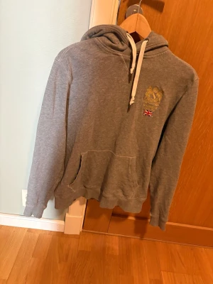 Grå hoodie med tryck och ficka - Grå hoodie med huva och vita dragsnören. Tryck med guldfärgad logga. Ett litet hål på bröstet som inte syns. Lite slutningar men dom är inte synliga. Bara där för modde. Storlek hade sagt mella s o xs 