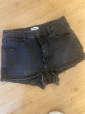 Svarta jeansshorts från Wrangler - Snygga svarta jeansshorts från Wrangler med råa kanter och klassisk femficksdesign. Shortsen har en normal passform och är perfekta för varma dagar. Stängs med knapp och dragkedja framtill.