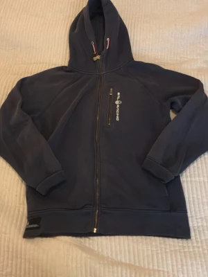 Marinblå hoodie från Sail Racing - Snygg marinblå hoodie från Sail Racing med dragkedja framtill och en bröstficka med zip. Hoodien har broderad logga på bröstet och muddar vid ärmslut och nederkant. Tillverkad i mjukt material och har en klassisk huva med dragsko.