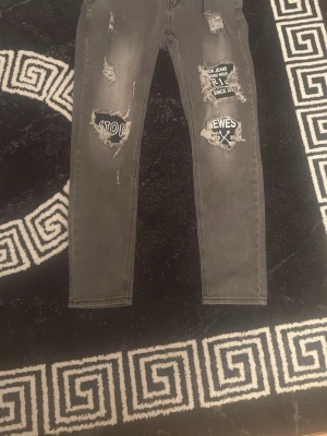 Gråa distressed jeans med tryck - Säljer ett par gråa jeans med slitningar och coola tryck på framsidan. Jeansen har skinny fit och flera trasiga detaljer med text som 'STOP' och 'JEWES'. Perfekta för dig som gillar en edgy streetstyle-look.