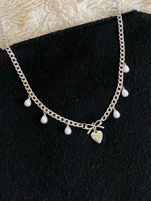 Silverfärgat halsband med pärlor och hjärta - Ett silverfärgat rostfritt halsband med kedja, dekorerat med små vita pärlor som hänger längs kedjan. I mitten finns en liten rosett och ett hjärtformat hänge. Halsbandet har en trendig och söt design, perfekt för att lyfta din outfit.