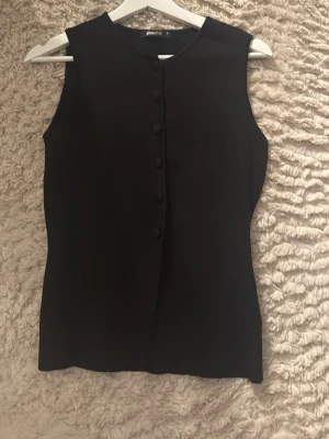 Svart topp med knappar från Gina Tricot - Stilren svart topp från Gina Tricot med rund halsringning och knäppning framtill. Toppen har en figurnära passform och är tillverkad i ett mjukt material som känns bekvämt mot huden. Använd endas 1-2 gånger så den är i jätte bra skick.