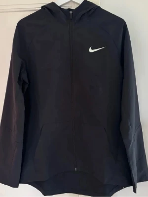 Svart Nike jacka med dragkedja - Svart hoodie från Nike med hel dragkedja framtill och vit Nike-logga på bröstet. Modellen har huva och två fickor framtill. Perfekt för en sportig och avslappnad stil. Materialet känns mjukt och följsamt.