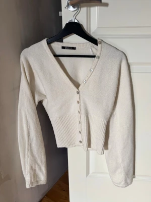 Vit ribbad kofta från Gina Tricot - Säljer en krämvit kofta från Gina Tricot med v-ringning och dekorativa pärlknappar framtill. Koftan har ribbad midja och långa ärmar, vilket ger en snygg och feminin siluett. Perfekt att styla med jeans eller kjol.