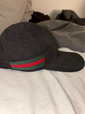 Svart Gucci keps med GG-mönster - Säljer en svart keps från Gucci med klassiskt GG-mönster över hela kepsen. På sidan finns en grön och röd rand som ger kepsen en ikonisk look. Materialet är bomull och kepsen har en böjd skärm. Perfekt för dig som vill ha en stilren accessoar. Storlek S ( 57 cm ) köpt från gucci. Skriv vid intrese! 