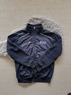 Svart vintage ziptröja från Hugo Boss - Snygg svart ziptröja från Hugo Boss med hög krage och dragkedja framtill. Tröjan har vita detaljer, två dragkedjefickor och broderad logga på bröstet och axeln. Perfekt för en stilren och sportig look.
