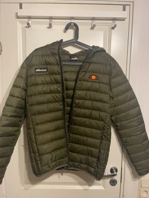 Olivgrön pufferjacka från Ellesse - Snygg olivgrön pufferjacka från Ellesse med huva och dragkedja framtill. Jackan har Ellesse-logga på bröstet och är quiltad för extra värme. Perfekt för kalla dagar och enkel att matcha med olika outfits.