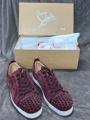 Vinröda Christian Louboutin sneakers - Snygga vinröda sneakers från Christian Louboutin i mocka med ikoniska nitar på tån och vita sulor. Skorna har låga skaft, matchande vinröda skosnören och exklusiv design som sticker ut. Perfekta för dig som vill ha något unikt och trendigt.
