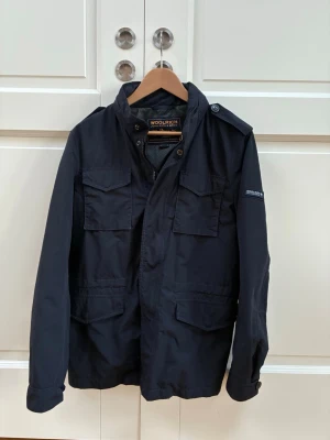  Field jacket Woolrich - Stilren svart vindjacka från Woolrich med fyra framfickor, axelklaffar och dragkedja med knappslå. Jackan har en klassisk krage och diskret logga på ärmen. Perfekt för blåsiga dagar och enkel att matcha med olika outfits. Jackan är i väldigt bra skick med nästan inga skavanker. Nypris är 6000 men säljer nu för 2000. Tveka inte över att höra av dig vid frågor eller prisförslag.