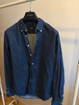 Blå denimskjorta från Morris - Säljer en klassisk blå denimskjorta från Morris med button down-krage och lång ärm. Skjortan har vita knappar och en liten röd broderad logga på bröstet. Perfekt till jeans eller chinos för en avslappnad men snygg look.