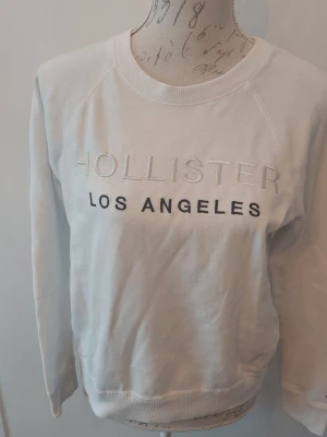 Vit sweatshirt från Hollister - Vit sweatshirt från Hollister med texten 'HOLLISTER LOS ANGELES' broderad på bröstet. Tröjan har rund halsringning, långa ärmar och ribbade muddar vid ärmslut och nederkant. 