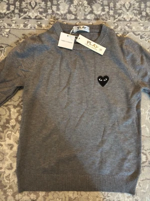Grå stickad tröja från Comme des Garçons - Snygg grå stickad tröja från Comme des Garçons PLAY med det ikoniska svarta hjärtat med ögon broderat på bröstet. Tröjan har rund halsringning och ribbade muddar vid ärmslut och nederkant. Perfekt för dig som gillar stilrena plagg med en twist.