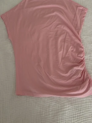 Rosa topp med rynkade detaljer 157 - Säljer en ljusrosa topp från 157 i mjukt och stretchigt material. Toppen har korta ärmar och en snygg rynkning på ena sidan som ger en cool och trendig look. Perfekt att matcha med jeans eller kjol för en avslappnad stil.