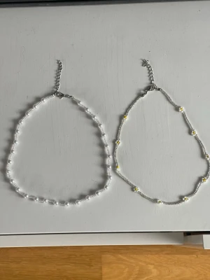 Två somriga halsband med kedja - Säljer två somriga halsband som jag köpt på Cypern . Det ena halsbandet har vita pärlor i olika storlekar och det andra har små vita pärlor med inslag av gula detaljer i form av prästkragar. Båda har silverfärgad kedja och justerbart lås. Perfekt accessoar för att lyfta din stil.