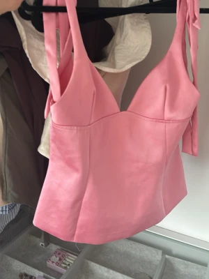Rosa korsettopp med knytband - Supersnygg rosa korsettopp med breda axelband som knyts på axlarna. Toppen har en figurnära passform och markerad byst, vilket ger en trendig siluett. Perfekt att styla med jeans eller kjol för en cool look.