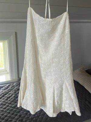 Vit sommarklänning med broderade blommor - Söt vit klänning med smala axelband och broderade blommor. Klänningen har en lätt utsvängd form och är tillverkad i ett luftigt material och öppen rygg. 