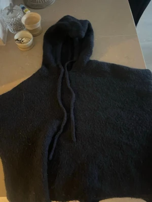 Svart stickad hoodie med huva - Mysig svart stickad hoodie med huva och dragsko. Oversized känsla och grovstickat material som ger en cool och avslappnad vibe. Perfekt för lager-på-lager och streetwear-stil.