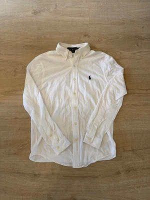 Vit skjorta från Ralph Lauren - Klassisk vit skjorta från Ralph Lauren med button-down krage och broderad logga på bröstet. Skjortan har långa ärmar, knappar framtill och är tillverkad i mjuk bomull. Perfekt för en clean och stilren look.