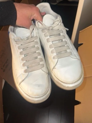 Vita Alexander McQueen sneakers - Stilrena vita sneakers från Alexander McQueen med blå mockadetalj och logga på hälen. Klassisk låg modell med bred sula och vita skosnören. Ovandel i läder och snygg kontrast mellan vitt och blått. Perfekta för en trendig streetstyle-look.