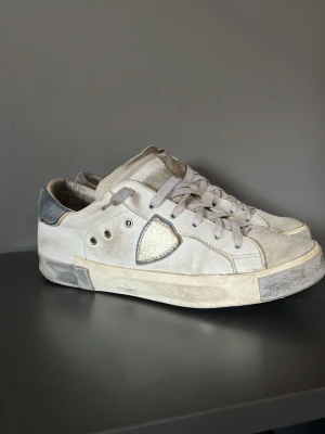 Vita sneakers från Philippe Model - Snygga vita sneakers från Philippe Model med grå detaljer och silverfärgad logga på sidan. Skorna har lågt skaft, vita skosnören och en slitstark sula. Ovandelen är i läder och mocka, med en cool vintage-look och kontrasterande hälparti. Om köpet skulle köpas vid instsbox skickar jag förmodligen samma dag som köpet☺️