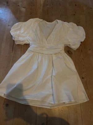Vit kort klänning med puffärmar - perfekt studentklänning  - Supersöt vit klänning med kort kjol, djup v-ringning och bred midjedetalj. Klänningen har puffiga korta ärmar och smock i ryggen för en bekväm passform. Perfekt för dig som gillar romantisk och trendig stil.