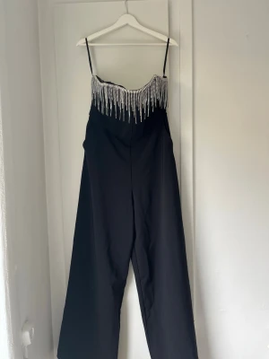 Svart jumpsuit med strassdetaljer - Supersnygg svart jumpsuit med smala axelband och vida ben. Överdelen har glittrande strassfransar som ger en riktigt festlig look. Perfekt för dig som vill sticka ut! Materialet känns mjukt och följsamt.