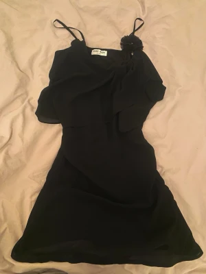 Svart kort klänning från Vero Moda - Säljer en svart kort klänning från Vero Moda med tunna axelband och en dekorativ blomdetalj vid axeln. Klänningen har ett lager av tyg som ger en snygg omlottkänsla och är perfekt för dig som gillar enkel men elegant stil.