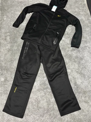 Svart Nike nocta tracksuit - Snygg svart tracksuit från Nike med hel dragkedja och huva. Jackan har två fickor med dragkedja och gul Nike-logga på bröstet. Byxorna har raka ben, fickor och gul Nike-text på ena benet. Tillverkad i ett mjukt, sportigt material som passar perfekt för chill eller träning.