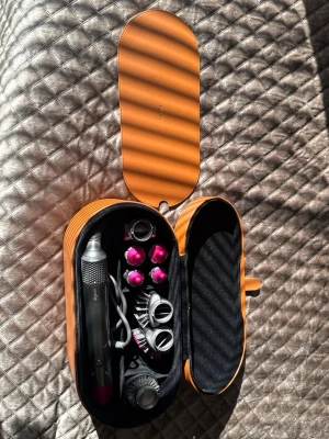 Dyson Airwrap multistyler med tillbehör - Säljer en Dyson Airwrap multistyler i snygg orange förvaringsväska. Inkluderar flera stylingtillbehör som borstar och locktillbehör i silver och lila. Perfekt för att skapa olika frisyrer med ett enda verktyg. Allt är snyggt organiserat i väskan.