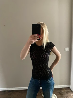 Svart spetsig topp med kort ärm - Snygg svart topp i spets med korta ärmar och tight passform. Toppen är genombruten och har ett feminint mönster som ger en cool look. Perfekt att styla med jeans eller kjol för en trendig outfit.