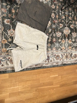 Grå Essentials shorts med snörning - Snygga ljusgrå shorts från Fear of God Essentials med svart logga på vänster ben. Shortsen har elastisk midja med snörning och är tillverkade i mjuk bomullsmix. Perfekta för en avslappnad och trendig look.