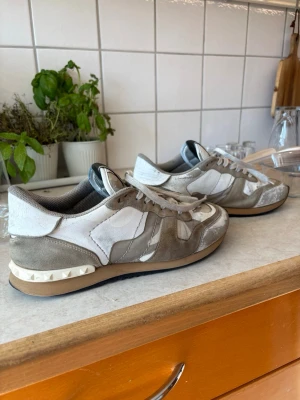 Valentino rockrunners - Snygga sneakers från Valentino i beige och vitt med detaljer i mocka och canvas. Skorna har en låg siluett, klassisk snörning och ikoniska nitar på hälen. Perfekta för dig som gillar exklusiva och trendiga sneakers med unik design. Inte toppskick, snygner som är lösa (går att fixa) och färg som släppt på tårna. Priset ej hugget i sten.