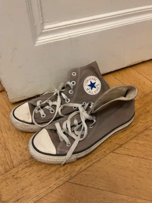Ljsubruna Converse All Star Highs - Klassiska grå Converse All Star high tops med vit tå och sula. Skorna har den ikoniska loggan på sidan, vita snören och gummisula med mönstrad undersida. Perfekta för en avslappnad och cool stil.