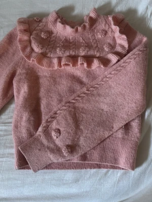 Rosa blus med volang och blommor - Jättefin rosa blus med volangdetaljer runt halsen och stickade blommor på bröstet och ärmsluten. Blusen har ribbade muddar och hög hals, vilket ger en gullig och trendig vibe. Materialet är mjukt och skönt, ser mer rosa ut i verkligheten än på bilden. 