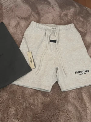 Essentials ljusgrå shorts med snörning - Ljusgrå shorts från Essentials Fear of God med snörning i midjan och logga tryckt på vänster ben. Mjukt material och relaxed fit, perfekta för sommaren. Klassisk design med elastisk midja och diskret svart detalj framtill. Helt NYA
