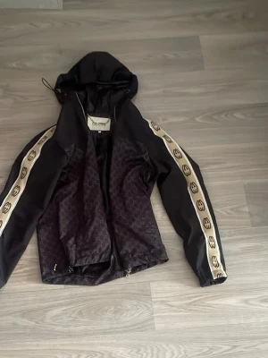Svart vindjacka från Gucci med huva - Svart vindjacka från Gucci med huva och dragkedja. Jackan har beige ränder med GG-logga längs ärmarna och diskret GG-mönster över hela tyget. Snygg streetstil och lätt material som passar perfekt till vardagslooken.