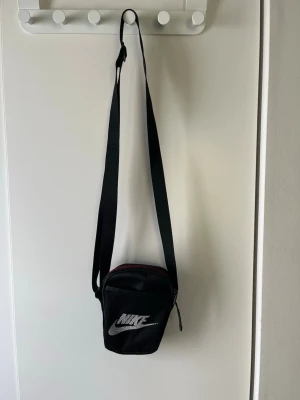 Svart axelväska från Nike - Snygg svart axelväska från Nike med justerbar rem och vit logga framtill. Väskan har dragkedja upptill och ett extra fack på baksidan. Perfekt storlek för mobil, plånbok och nycklar. Tillverkad i slitstarkt syntetmaterial.